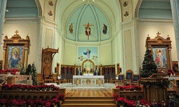 An elaborately decorated church altar with a crucifix centerpiece. Floral arrangements and Christmas trees adorn the sides. Two large religious paintings flank the altar, adding to the festive ambianc