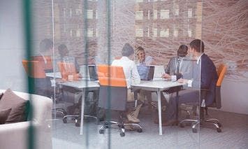 People seated at a conference table working on laptops inside a modern office with glass walls, orange and white chairs, and a large abstract wall art.
