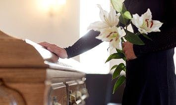 A person dressed in black holds white lilies, placing a hand on a wooden casket inside a softly lit room.