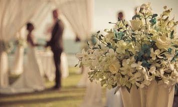 A bouquet of white flowers sits in the foreground while a couple stands and exchanges vows under a draped canopy in an outdoor wedding setting.